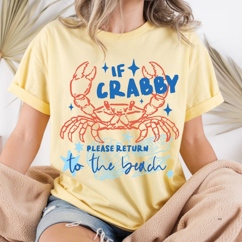 Puede incluir: Camiseta amarilla clara con un dise&ntilde;o de cangrejo rojo y azul. El texto dice "If Crabby Please Return to the Beach". La camiseta tiene mangas enrolladas y un corte holgado.
