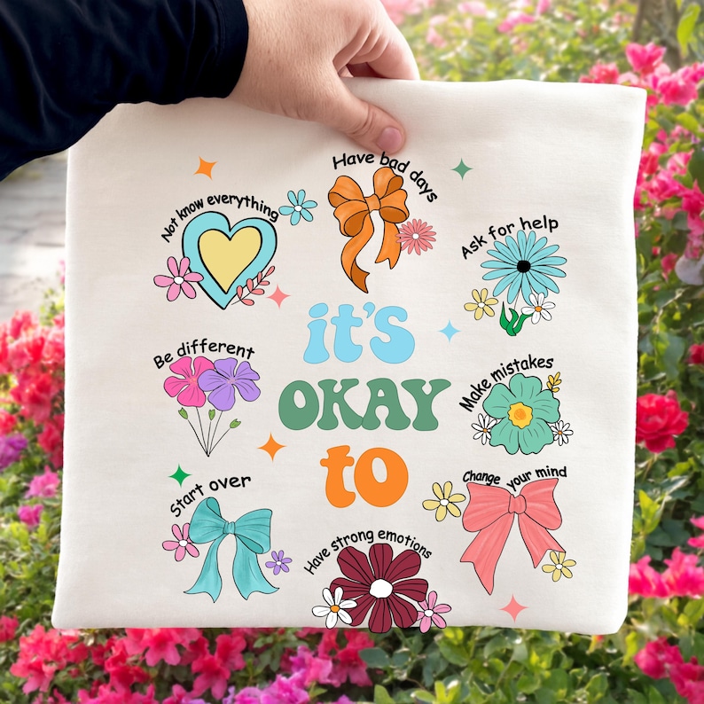 May include: Cream-colored tote bag with the words "it's OKAY to" in a retro font, surrounded by colorful illustrations and phrases like "Have bad days" and "Ask for help." The design includes flowers, bows, and stars.