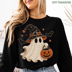 May include: A black long-sleeve sweatshirt featuring a graphic of a white ghost with black polka dots wearing a black and orange plaid witch hat. The ghost holds an orange jack-o'-lantern bucket, surrounded by black bats and orange stars.