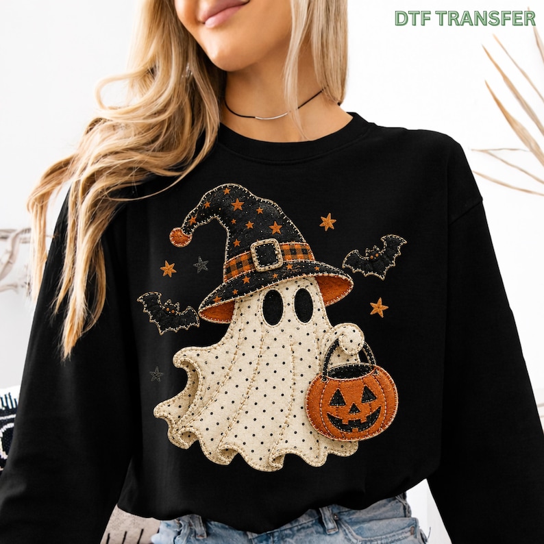 May include: A black long-sleeve sweatshirt featuring a graphic of a white ghost with black polka dots wearing a black and orange plaid witch hat. The ghost holds an orange jack-o'-lantern bucket, surrounded by black bats and orange stars.
