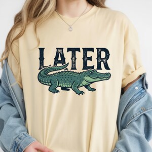 May include: A light yellow t-shirt featuring a green alligator illustration below the word "LATER" in dark blue, distressed font. The alligator is detailed with scales and a smiling expression.