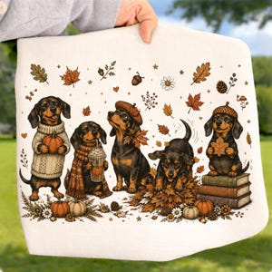 May include: A white fabric item featuring a colorful illustration of five dachshunds in autumn settings. The dogs are depicted with pumpkins, coffee cups, fall leaves, acorns, and books, surrounded by small stars, flowers, and seasonal foliage.