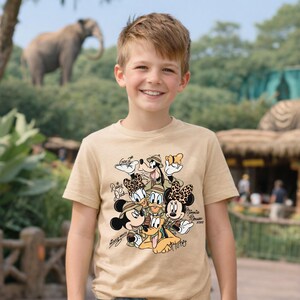 Puede incluir: Una camiseta color beige con un dise&ntilde;o de personajes de Disney vestidos para un safari. Los personajes incluyen Mickey Mouse, Minnie Mouse, Donald Duck, Daisy Duck, Goofy y Pluto. La camiseta la lleva un ni&ntilde;o sonriente.