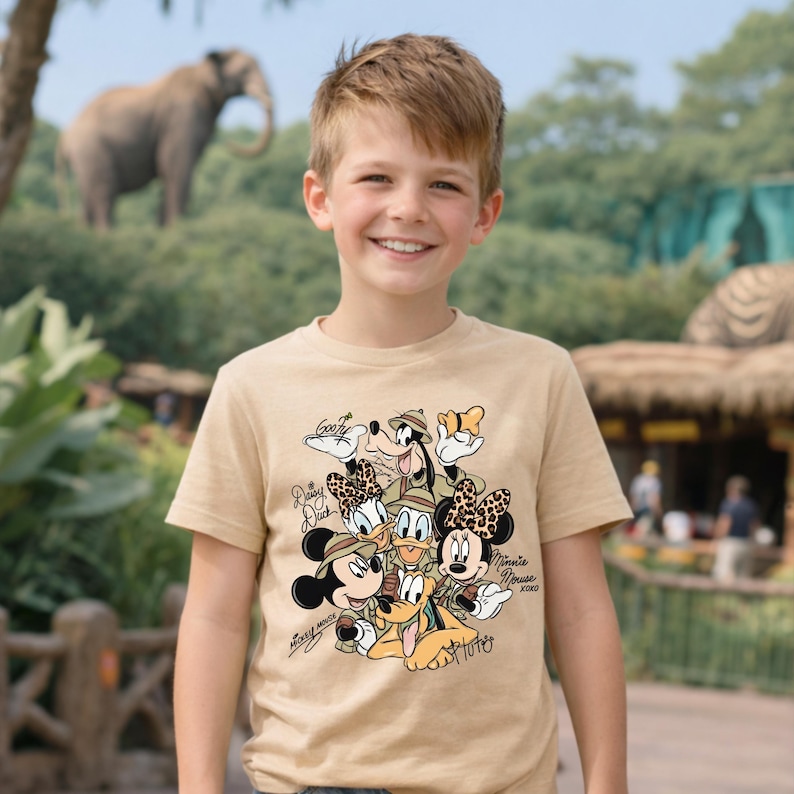 Puede incluir: Una camiseta color beige con un dise&ntilde;o de personajes de Disney vestidos para un safari. Los personajes incluyen Mickey Mouse, Minnie Mouse, Donald Duck, Daisy Duck, Goofy y Pluto. La camiseta la lleva un ni&ntilde;o sonriente.