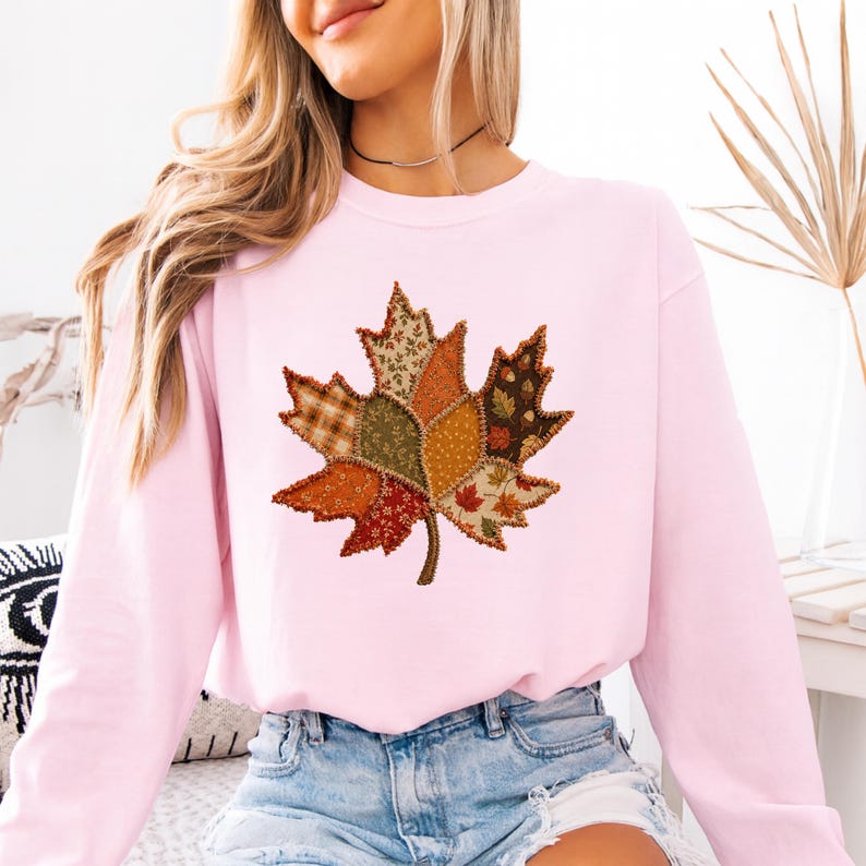 May include: A light pink long-sleeve sweatshirt featuring a large central graphic of a maple leaf composed of various patchwork-style fabric patterns in shades of orange, brown, green, and cream with visible stitched edges throughout the design.