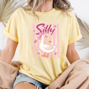 May include: Yellow t-shirt featuring a cartoon goose with a pink bow and the word "Silly". The goose is set against a pink and white striped background with star accents. The shirt has rolled-up sleeves.