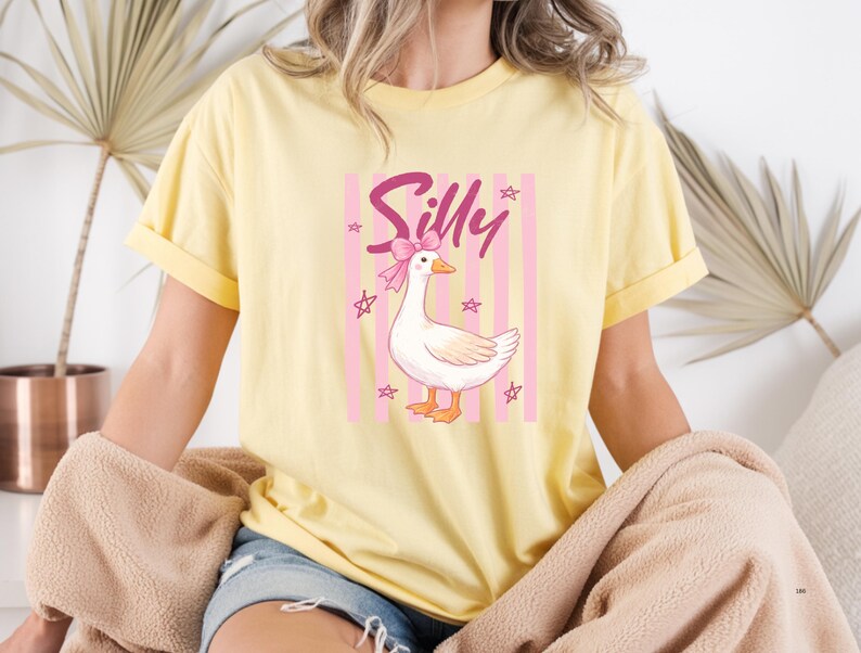 May include: Yellow t-shirt featuring a cartoon goose with a pink bow and the word "Silly". The goose is set against a pink and white striped background with star accents. The shirt has rolled-up sleeves.