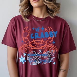 Puede incluir: Camiseta burdeos con un dise&ntilde;o de cangrejo y conchas marinas. El cangrejo naranja tiene el texto azul "IF CRABBY" encima. Debajo, conchas marinas azules y blancas y el texto "PLEASE BE".