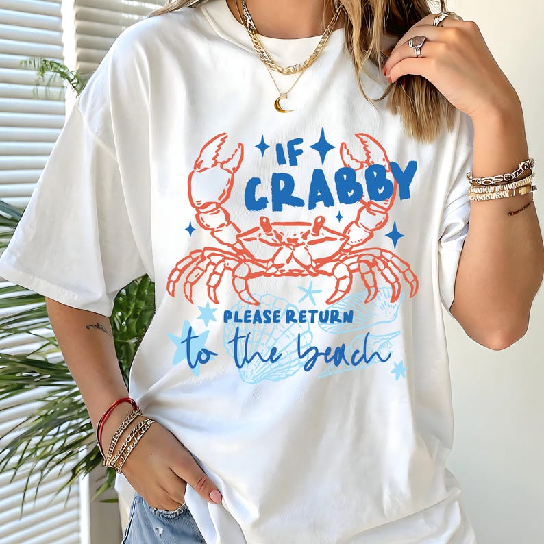 Puede incluir: Camiseta blanca con un dise&ntilde;o de cangrejo en rojo y azul, y el texto "If Crabby Please Return to the Beach". El dise&ntilde;o incluye estrellas y una ilustraci&oacute;n de concha. La camiseta tiene mangas cortas.