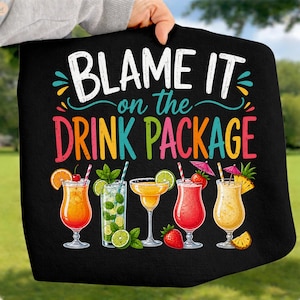 May include: Black towel featuring the phrase "BLAME IT on the DRINK PACKAGE" in white, pink, and green. The towel displays illustrations of five colorful cocktails with garnishes like oranges, limes, strawberries, and pineapple.
