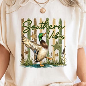 May include: Cream-colored t-shirt featuring a graphic of a duck with outstretched wings, stars, and the text "Southern Vibes" in green script. The design includes green and brown vertical stripes.