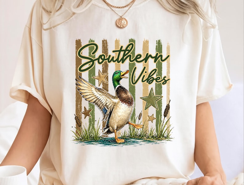 May include: Cream-colored t-shirt featuring a graphic of a duck with outstretched wings, stars, and the text "Southern Vibes" in green script. The design includes green and brown vertical stripes.