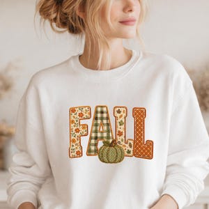 May include: A white crewneck sweatshirt featuring the word FALL in large block letters. Each letter has a unique pattern: floral, green and white gingham, autumn leaves, and orange polka dots. A green pumpkin sits below the letter A.