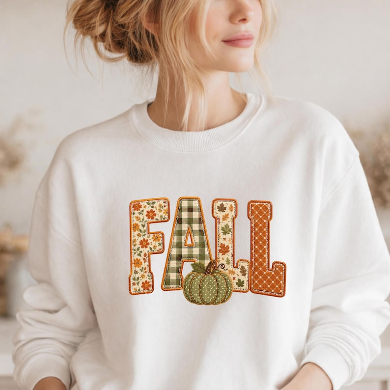May include: A white crewneck sweatshirt featuring the word FALL in large block letters. Each letter has a unique pattern: floral, green and white gingham, autumn leaves, and orange polka dots. A green pumpkin sits below the letter A.