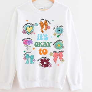 May include: White sweatshirt with a colorful graphic design. The design includes the phrase "it's OKAY to" with various positive affirmations and floral and bow illustrations. The sweatshirt is hanging on a wooden hanger.