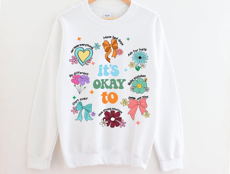 May include: White sweatshirt with a colorful graphic design. The design includes the phrase "it's OKAY to" with various positive affirmations and floral and bow illustrations. The sweatshirt is hanging on a wooden hanger.