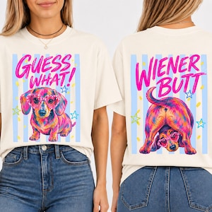 May include: Cream-colored t-shirt with a colorful dachshund wearing heart-shaped glasses. The front of the shirt says "GUESS WHAT!" in pink, while the back says "WIENER BUTT" in pink. Blue and white stripes and stars decorate the design.