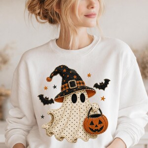 May include: A white crewneck sweatshirt featuring a central graphic of a cream-colored ghost with black polka dots. The ghost wears a black witch hat with orange stars and a plaid band, and carries an orange jack-o'-lantern bucket with black bats.