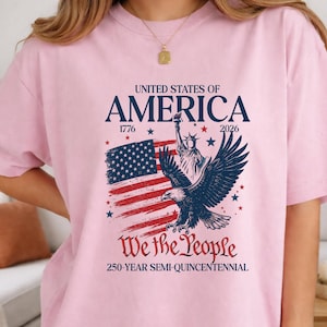 May include: A light pink t-shirt featuring a patriotic design. The graphic includes the words "UNITED STATES OF AMERICA" above an image of the American flag, the Statue of Liberty, and an eagle. The text "We the People" and "250-YEAR SEMI-QUINCENTENNIAL" are also included.