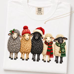May include: A white t-shirt featuring a graphic of five sheep standing in a row. The sheep wear festive winter accessories including a red pom-pom hat, a Santa hat, a red and green striped scarf, and a green patterned scarf with a small bell.