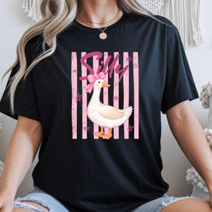 May include: Black t-shirt featuring a white goose with a pink bow and the word "Silly" in pink script. The goose is set against a pink striped background with star accents. A casual, fun design.
