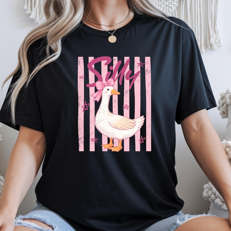 May include: Black t-shirt featuring a white goose with a pink bow and the word "Silly" in pink script. The goose is set against a pink striped background with star accents. A casual, fun design.