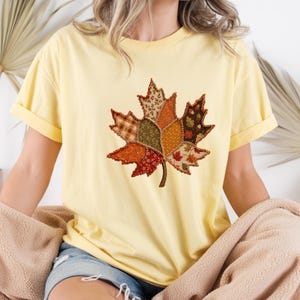 May include: A pale yellow short-sleeve t-shirt featuring a large central graphic of a maple leaf composed of various patchwork fabric patterns in shades of orange, brown, green, and cream with visible dark brown stitched outlines.