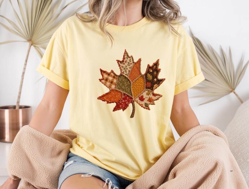 May include: A pale yellow short-sleeve t-shirt featuring a large central graphic of a maple leaf composed of various patchwork fabric patterns in shades of orange, brown, green, and cream with visible dark brown stitched outlines.