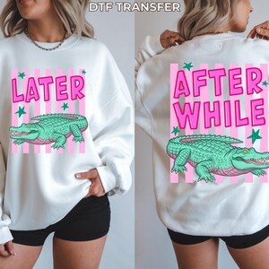 May include: White sweatshirt with a pink and white striped design. The front features the word "LATER" and a green alligator graphic. The back says "AFTER WHILE" with a matching alligator graphic. The text "DTF TRANSFER" is at the top.