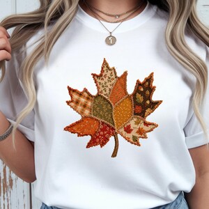 May include: A white short-sleeved t-shirt featuring a large central graphic of a maple leaf. The leaf is composed of various patchwork-style fabric patterns in shades of orange, brown, green, and yellow, with visible frayed edges.