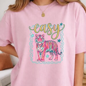 May include: Pink t-shirt featuring a colorful tiger illustration with the word "easy" in a cursive font. The tiger has stripes of pink, blue, and yellow. The design is framed with a light blue border and stars.