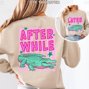 May include: Beige sweatshirt with a pink and white striped design. The back features the text "AFTER WHILE" in pink, with a green alligator graphic. The front has a smaller image with the word "LATER" and an alligator.