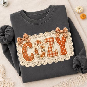 May include: A charcoal gray crewneck sweatshirt featuring a central rectangular patch with a cream-colored scalloped lace border. The patch displays the word COZY in orange fabric letters with floral, plaid, and polka dot patterns and two plaid bows.