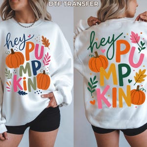 May include: A white long-sleeve sweatshirt featuring the text "hey PUMPKIN" in colorful letters, accompanied by orange pumpkin illustrations and various autumn-themed leaf graphics in shades of pink, green, brown, and yellow, with "DTF TRANSFER" text.