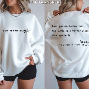 May include: A white crewneck sweatshirt featuring black text. The front displays the phrase 'you are enough'. The back displays the text 'Dear person behind me, the world is a better place with you in it love, the person in front of you'.