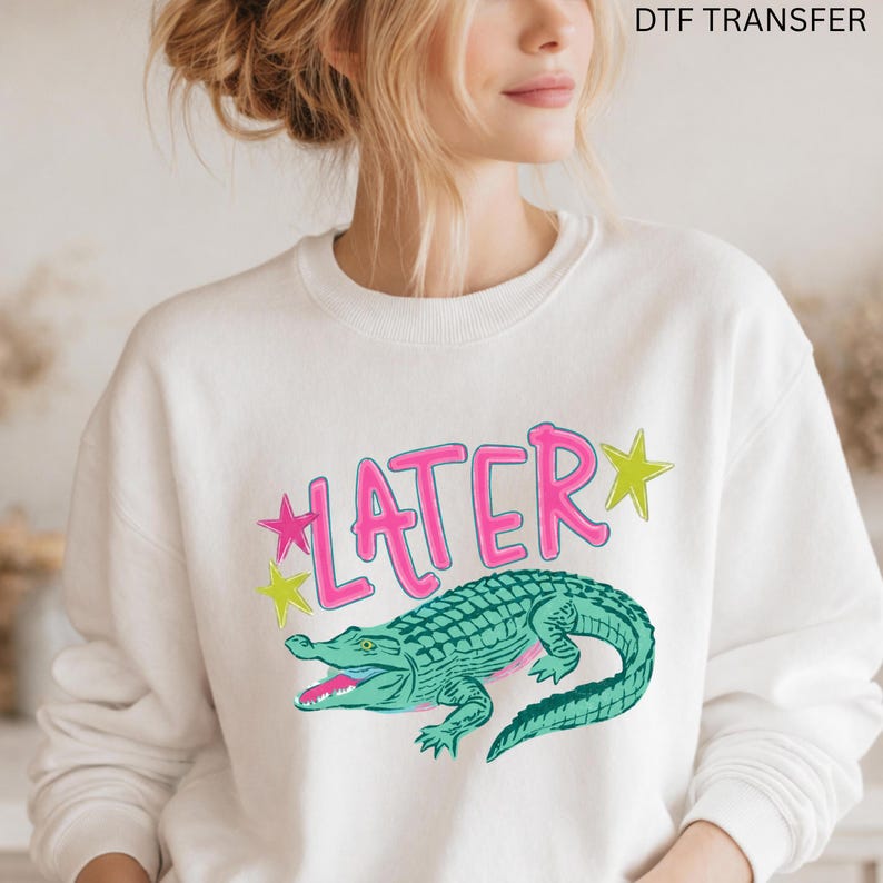 May include: White sweatshirt with a graphic of a green alligator and the word "LATER" in pink with yellow star accents. The text "DTF TRANSFER" is at the top.