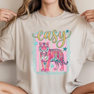 May include: Beige t-shirt featuring a colorful tiger illustration with pink, blue, and yellow stripes. The word "easy" is written in cursive above the tiger, with stars and a decorative border.