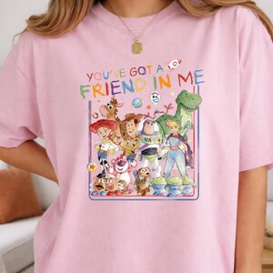 K&ouml;nnte beinhalten: Ein hellrosa T-Shirt mit einer bunten Grafik verschiedener animierter Spielzeugfiguren. Der Text "YOU'VE GOT A FRIEND IN ME" ist in mehrfarbigen Buchstaben &uuml;ber den Figuren gedruckt, darunter ein Cowboy, ein Weltraum-Ranger und andere.
