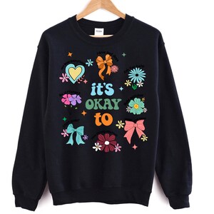May include: Black sweatshirt with a colorful graphic design. The design includes flowers, bows, hearts, and the phrase "it's okay to." The sweatshirt is hanging on a wooden hanger against a white background.