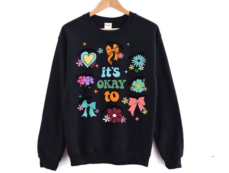 May include: Black sweatshirt with a colorful graphic design. The design includes flowers, bows, hearts, and the phrase "it's okay to." The sweatshirt is hanging on a wooden hanger against a white background.