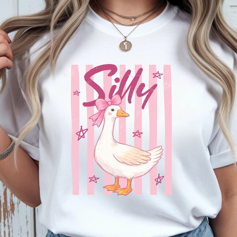 May include: White t-shirt featuring a cartoon goose with a pink bow and the word "Silly" in pink script. The design includes pink vertical stripes and star accents, creating a playful and whimsical aesthetic.