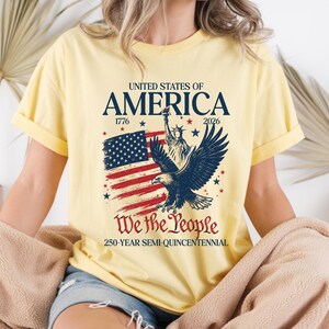 May include: A light yellow t-shirt featuring a patriotic design. The graphic includes the text "UNITED STATES OF AMERICA" with the years 1776 and 2026, an American flag, an eagle, and the words "We the People".