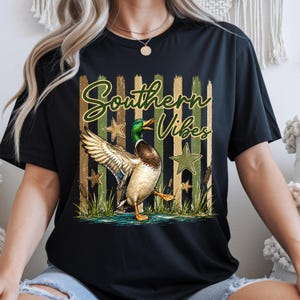 May include: Black t-shirt with a graphic of a mallard duck with outstretched wings, the text "Southern Vibes", and star accents. The duck is depicted in a naturalistic style with green, brown, and white feathers. The background features vertical stripes.