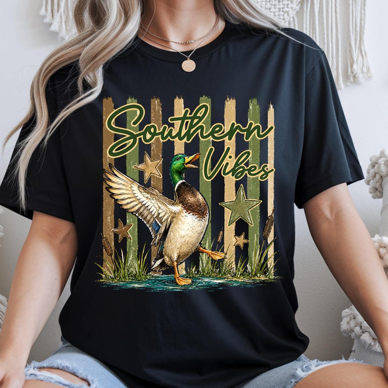May include: Black t-shirt with a graphic of a mallard duck with outstretched wings, the text "Southern Vibes", and star accents. The duck is depicted in a naturalistic style with green, brown, and white feathers. The background features vertical stripes.
