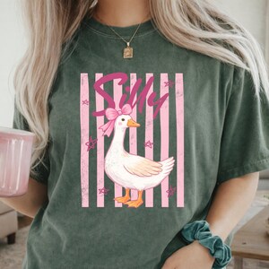 May include: Dark green t-shirt featuring a cartoon goose with a pink bow and the word "Silly" in pink script. The goose is set against a backdrop of pink and white stripes and star shapes.