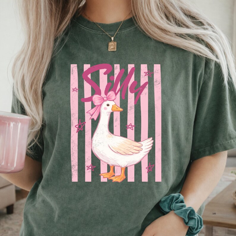 May include: Dark green t-shirt featuring a cartoon goose with a pink bow and the word "Silly" in pink script. The goose is set against a backdrop of pink and white stripes and star shapes.