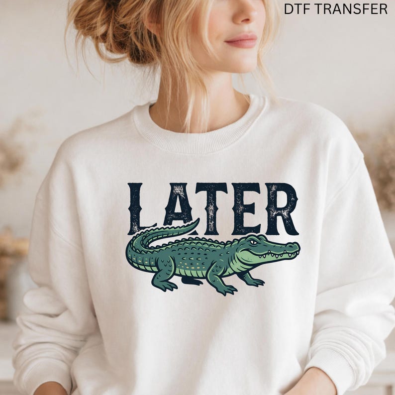 May include: White sweatshirt with the word "LATER" in dark blue, above a green alligator illustration. The sweatshirt is a crew neck style. The text "DTF TRANSFER" is at the top right.