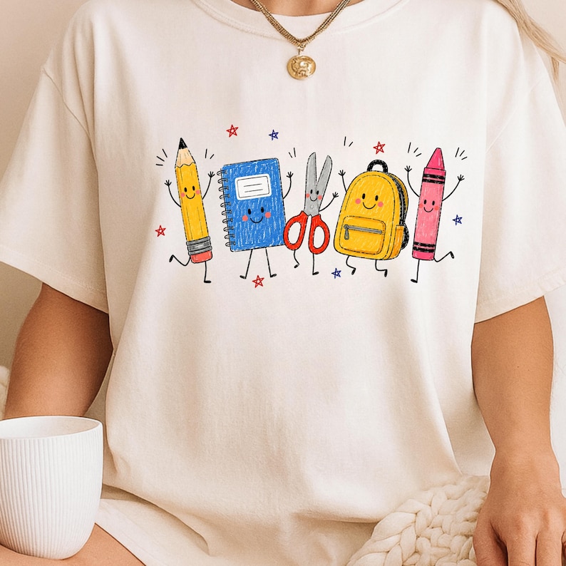 May include: A cream-colored t-shirt featuring a graphic print of five anthropomorphic school supplies: a yellow pencil, a blue notebook, red scissors, a yellow backpack, and a pink crayon, all accented with small red and blue stars.