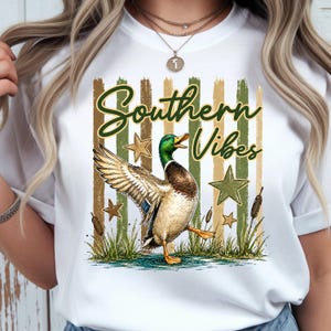 May include: White t-shirt featuring a graphic of a mallard duck with the text "Southern Vibes." The design includes green and brown stripes, stars, and cattails. The duck is depicted in a pond.
