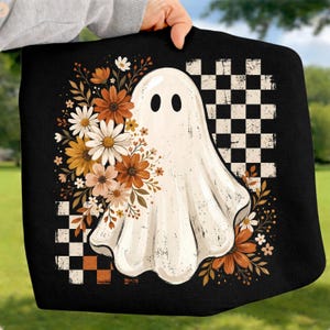 May include: A black fabric item featuring a central white ghost graphic surrounded by a cluster of orange, brown, and white flowers. The design includes a distressed black and white checkered pattern in the background corners of the illustration.
